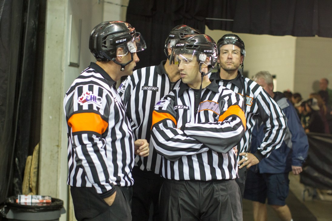 Three hockey referees in black and white striped jerseys with helmets gathered together before a game