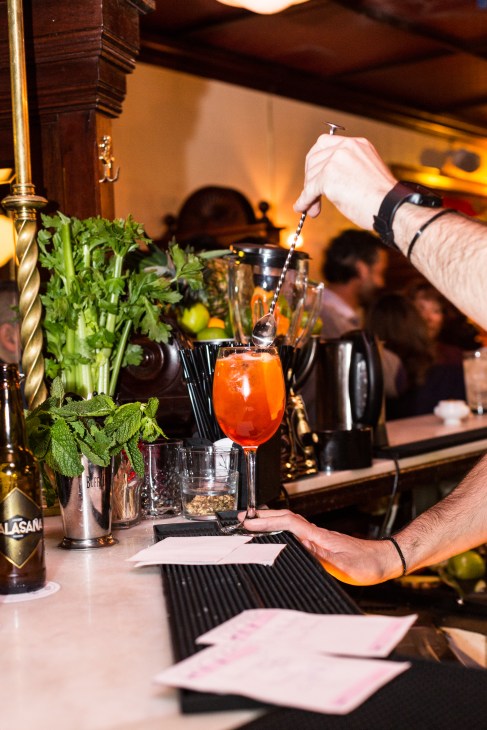 Bartender pouring liquid into a red cocktail at a bar with fresh herbs and bottles visible.