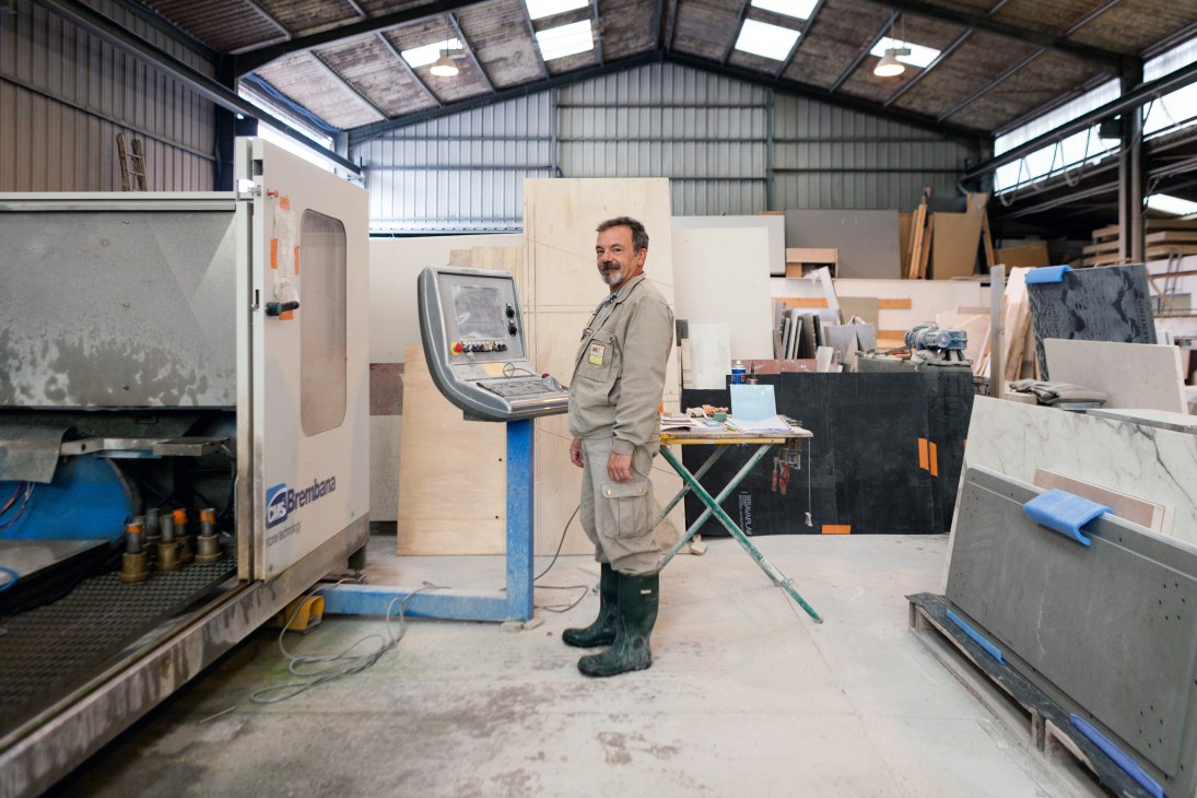 Worker in beige overalls stands at a CNC control panel in a marble factory workshop.