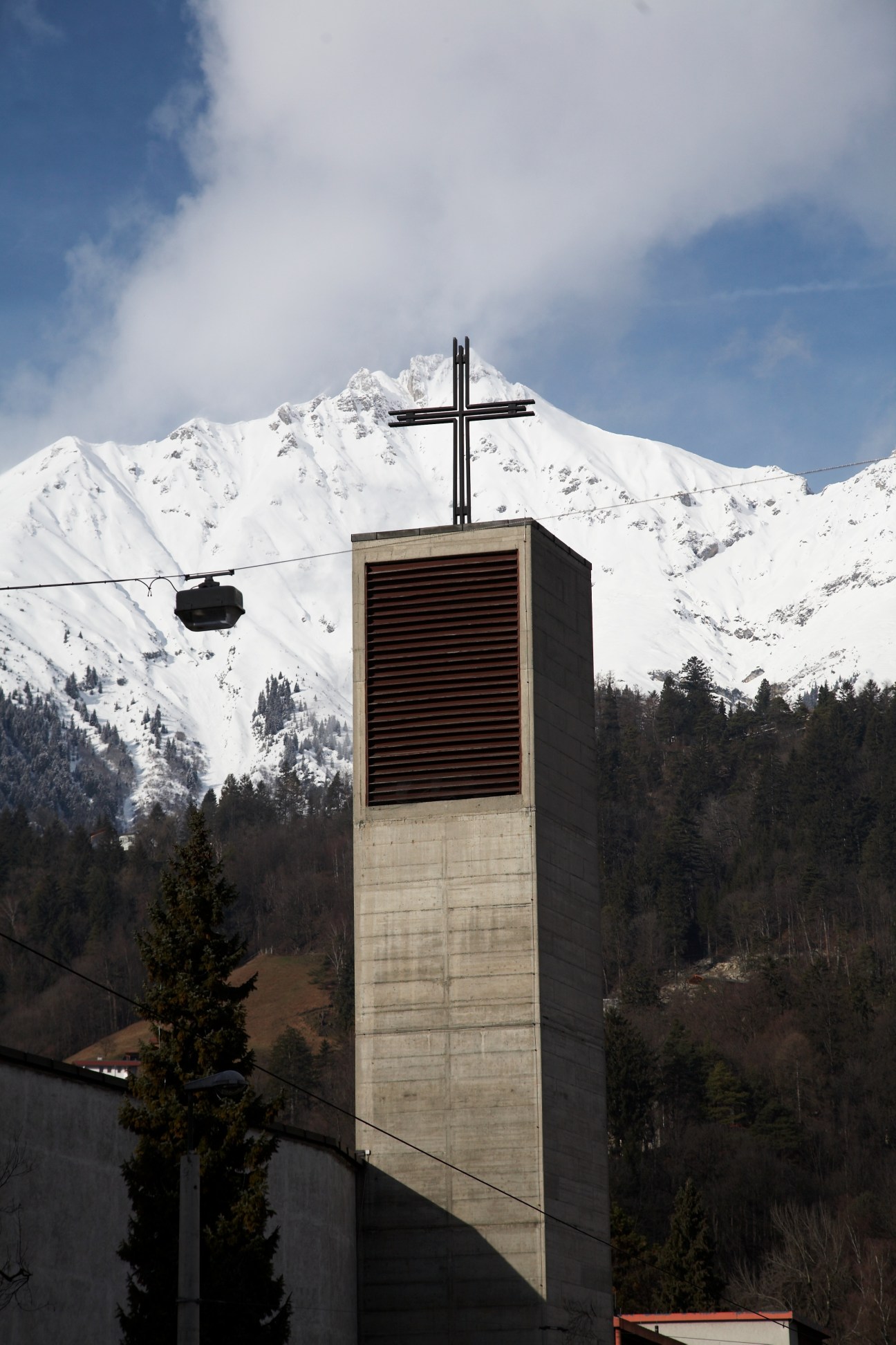 Modernist concrete church tower with black cross against snow-covered Alpine mountains in Saggen, Innsbruck.