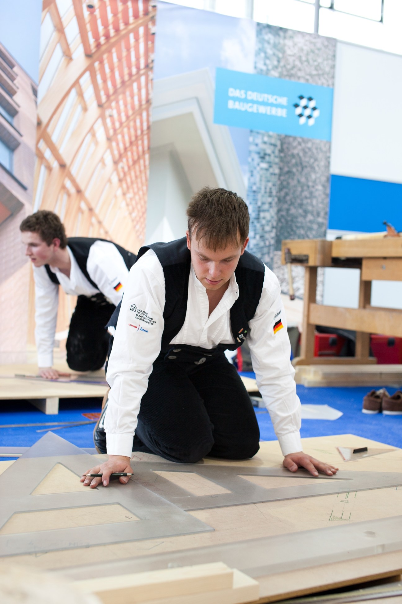Apprentice carpenter in traditional white shirt and black trousers works on wood flooring at BAU fair.