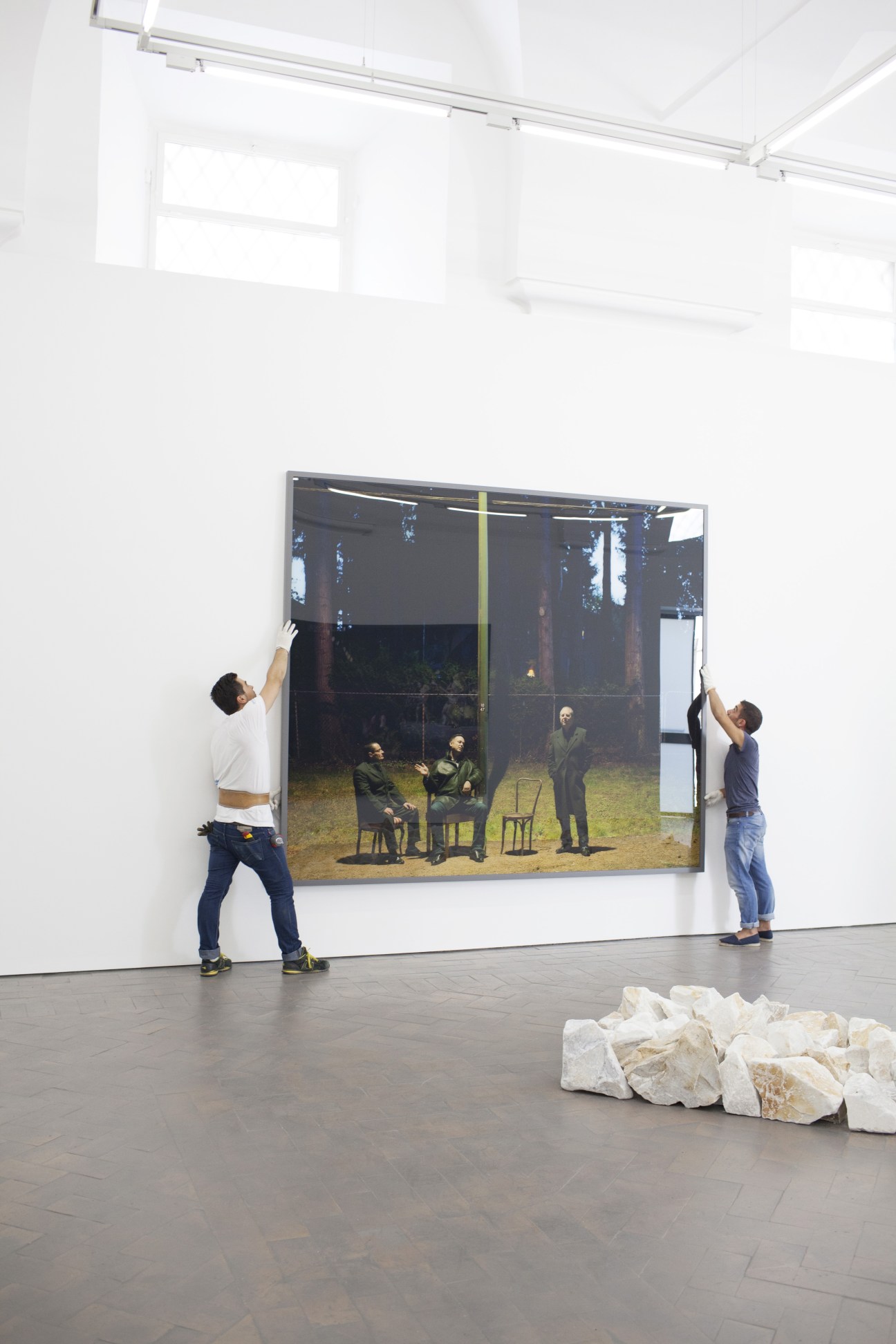 Two technicians hang a large framed Jeff Wall photograph on a white gallery wall.