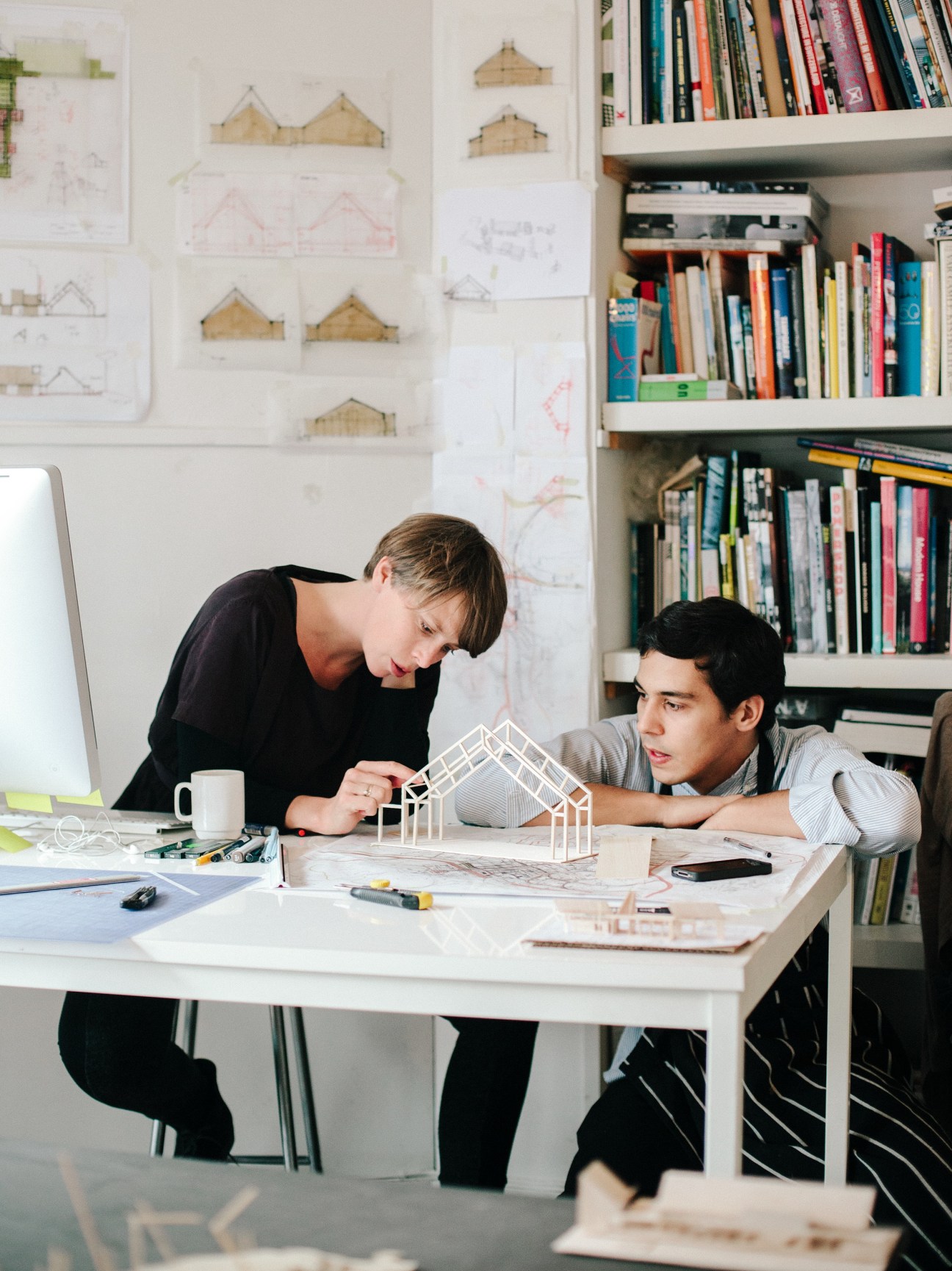Two architects review a white architectural model at a desk in a studio with sketches and books.