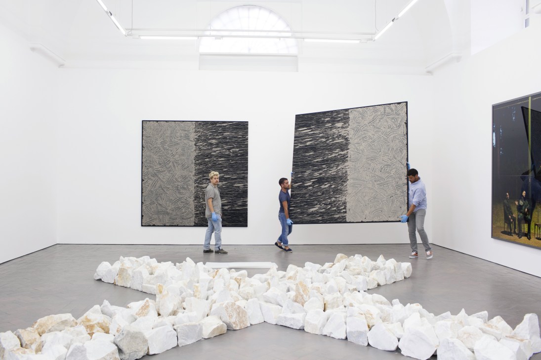 Three technicians position artworks in a white gallery space with high ceilings and a Richard Long stone installation in the foreground.
