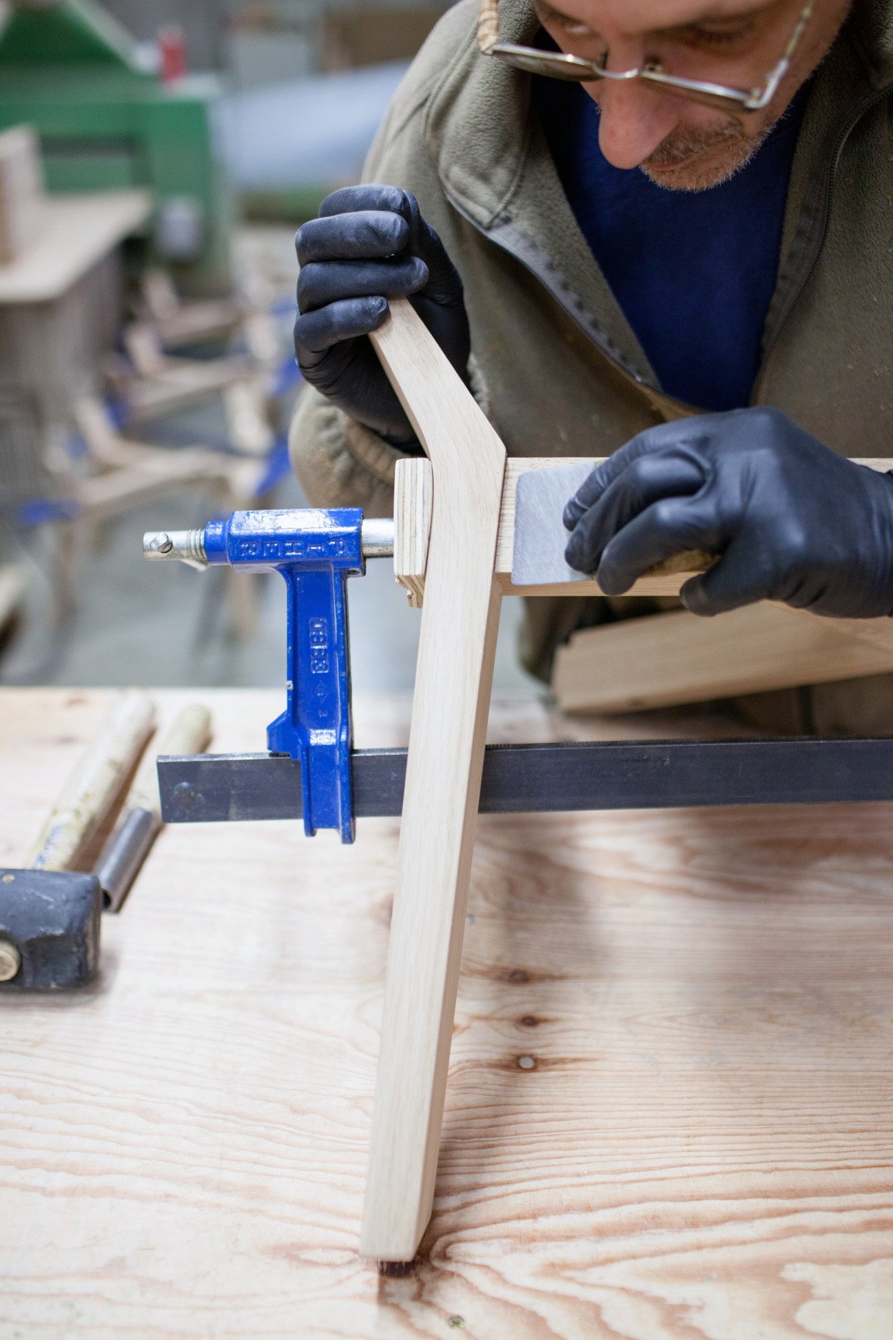 Craftsman in glasses and dark gloves uses a blue clamp to secure light wood pieces during furniture assembly at Alki factory.