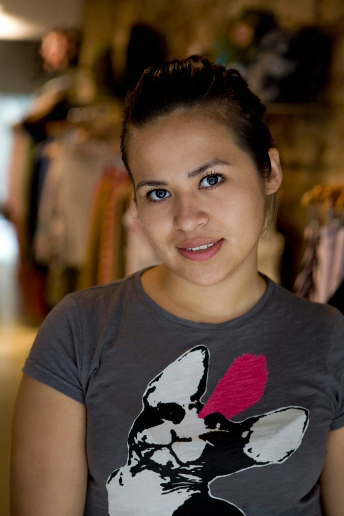 Portrait of Joselin Sanchez, a woman with dark hair styled in an updo, wearing a graphic t-shirt with a dog design, smiling at the camera in what appears to be a clothing boutique.