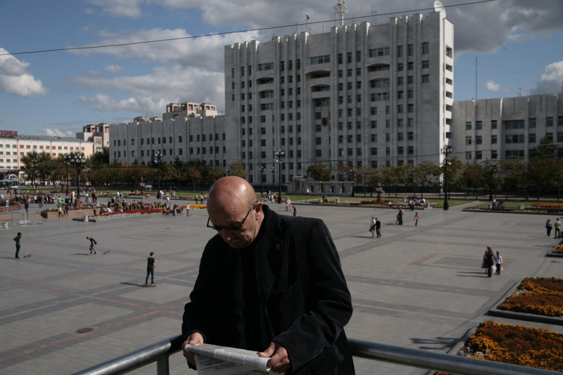 Man in black coat reading newspaper overlooking Lenin Square with Soviet-era apartment buildings.