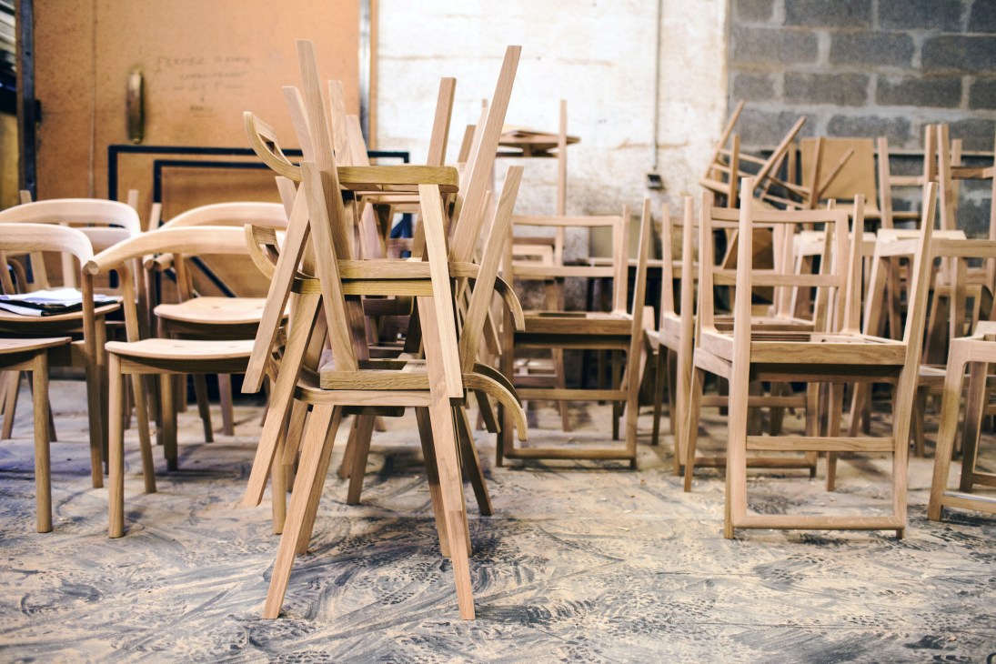 Wooden chair frames and partially assembled furniture pieces in a factory workshop with brick walls.