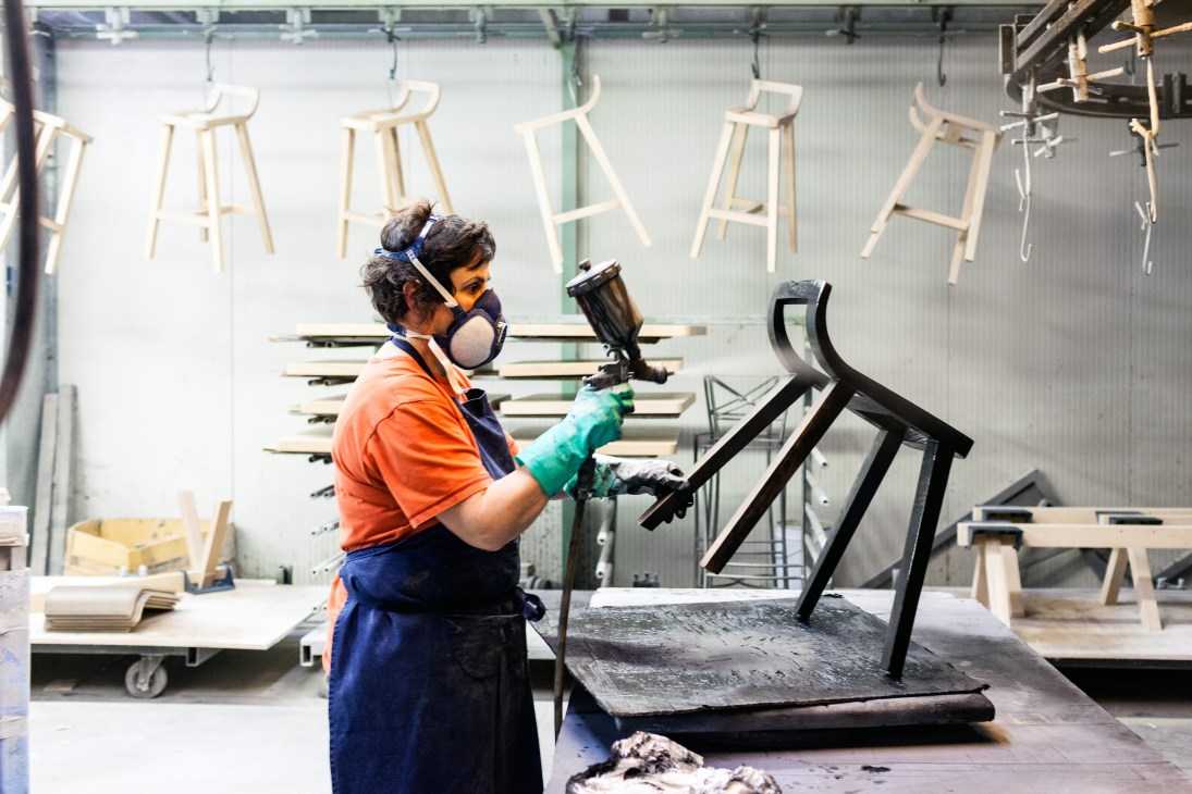 Factory worker in orange shirt painting a black chair frame in an industrial workshop.