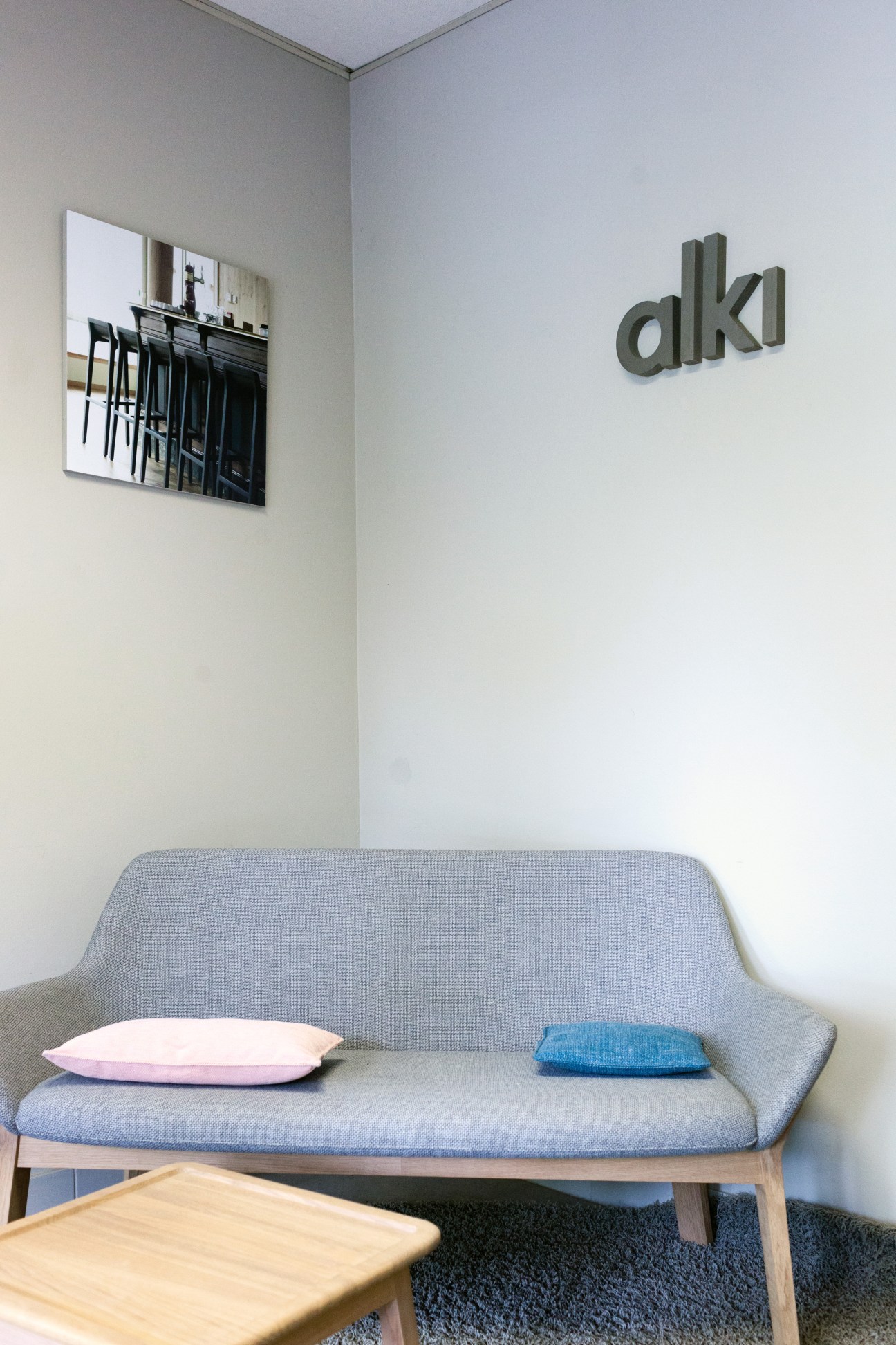 Alki furniture showroom displaying a grey upholstered sofa with wooden frame, pink and blue pillows, and the Alki logo on the wall above.