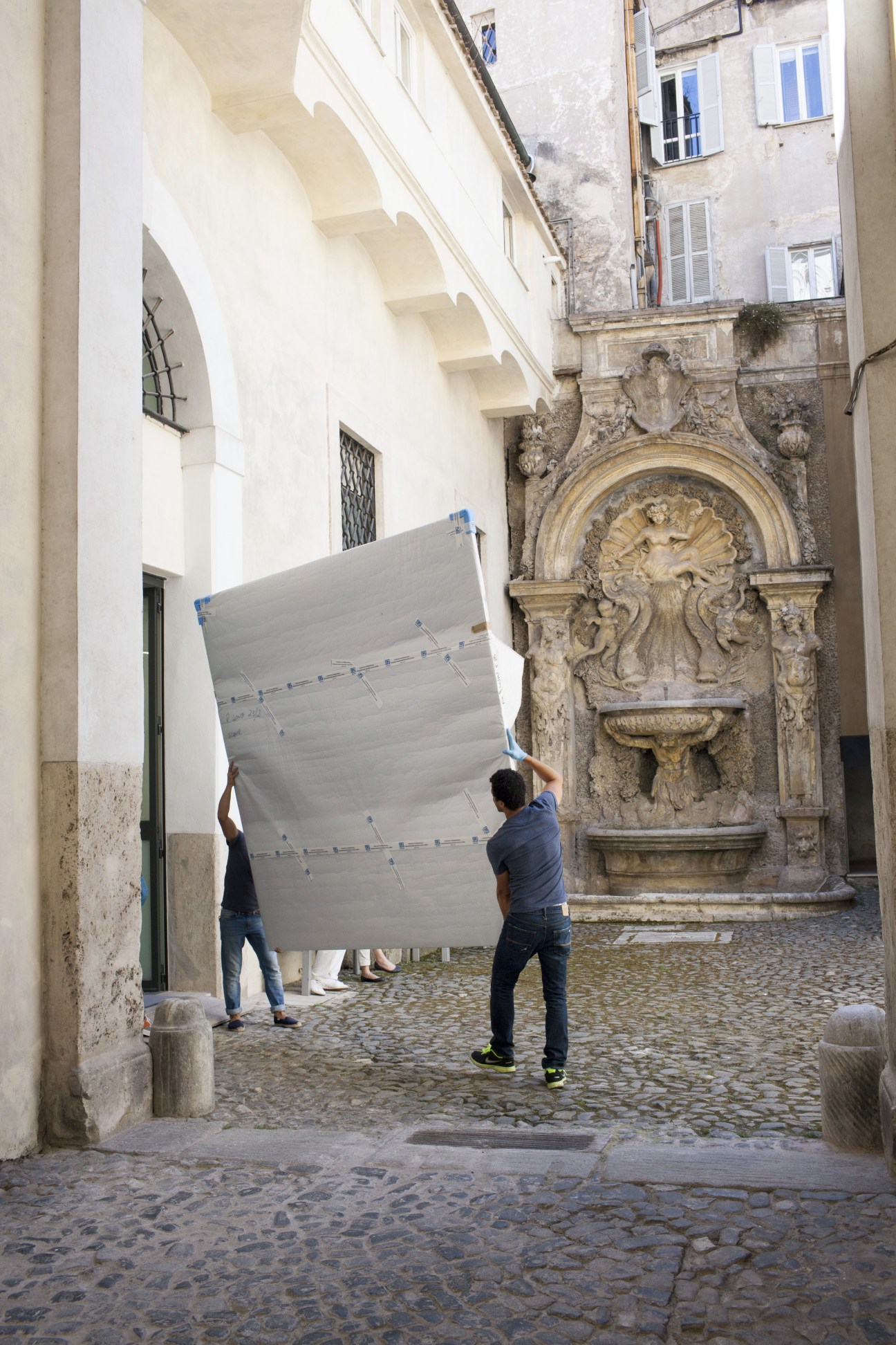 Two technicians carry a large framed artwork through a Roman courtyard with baroque architectural details.