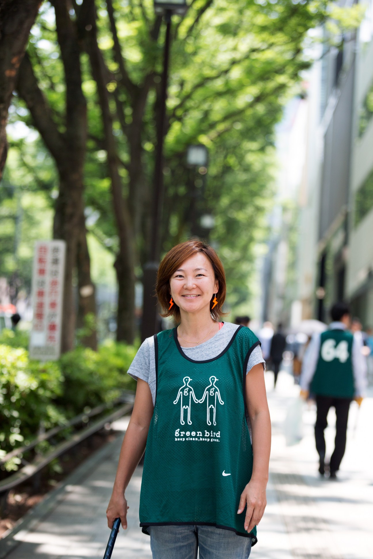 Mika Arai, Omotesando Green Bird team leader, wearing a green Nike bib with the Green Bird logo on a tree-lined Tokyo street.