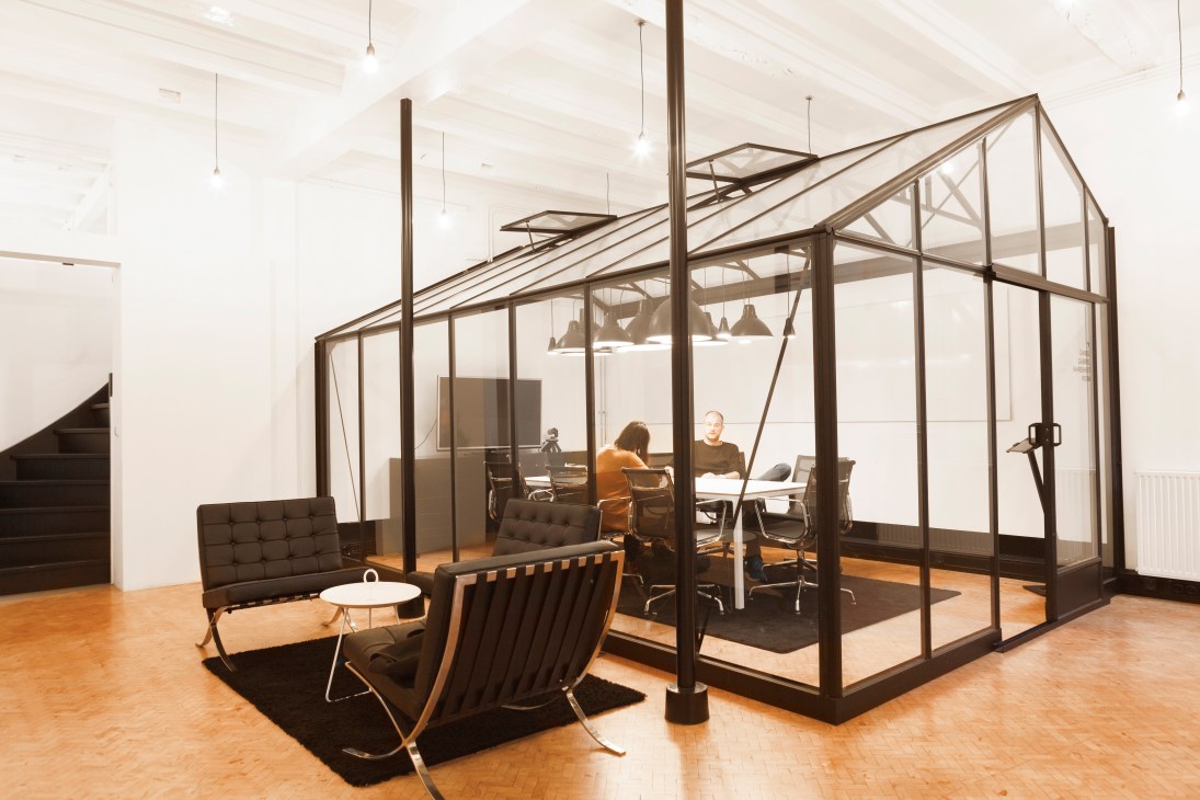 Modern shared office workspace with glass-partitioned meeting rooms and lounge seating on wooden flooring.