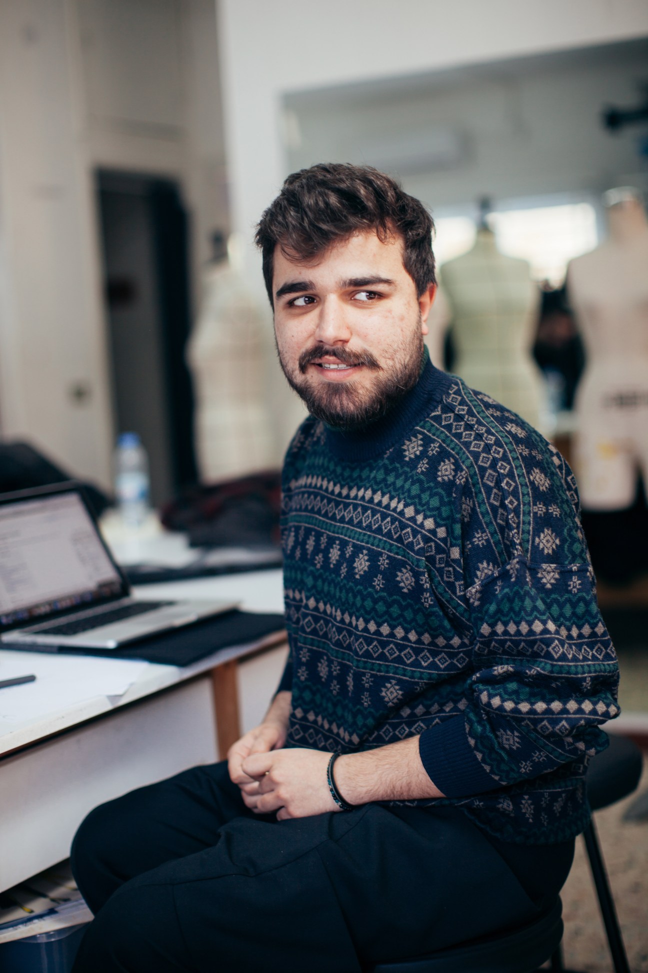 A male student wearing a patterned sweater sits at a desk in a design studio workspace.