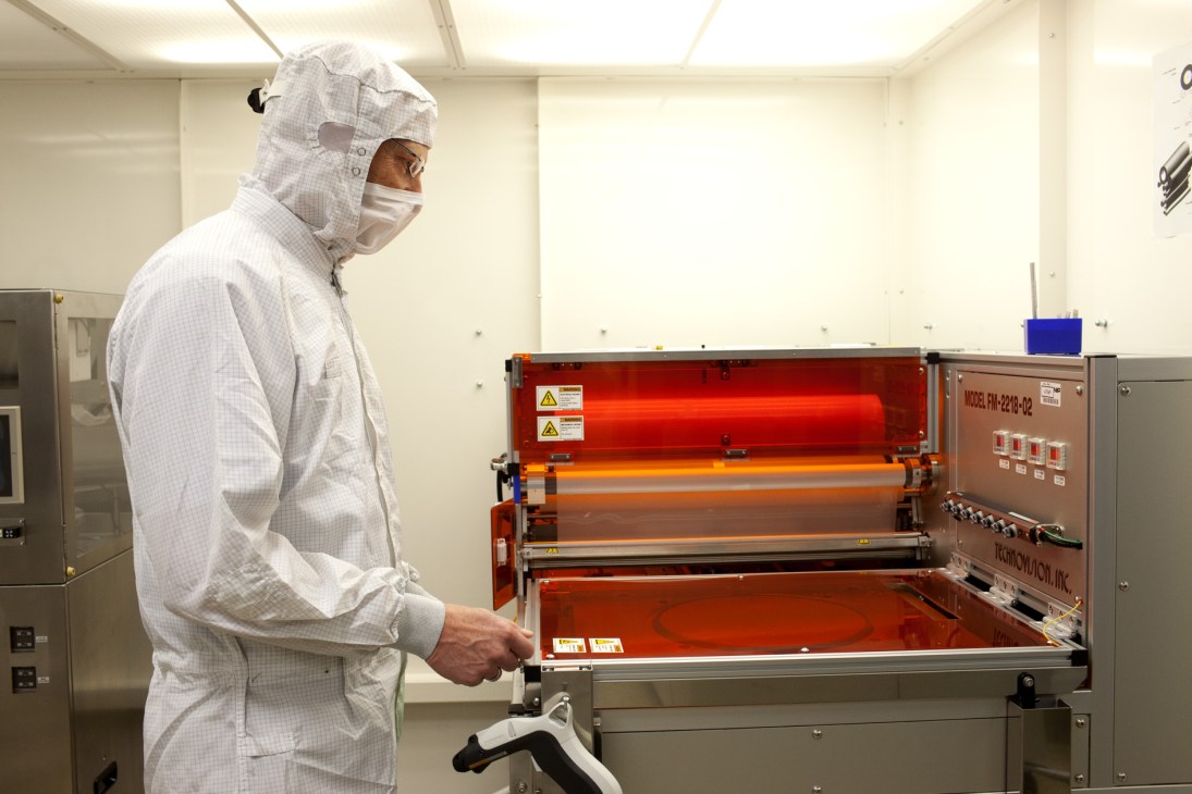 Worker in cleanroom suit operating semiconductor wafer production equipment with red heating elements.
