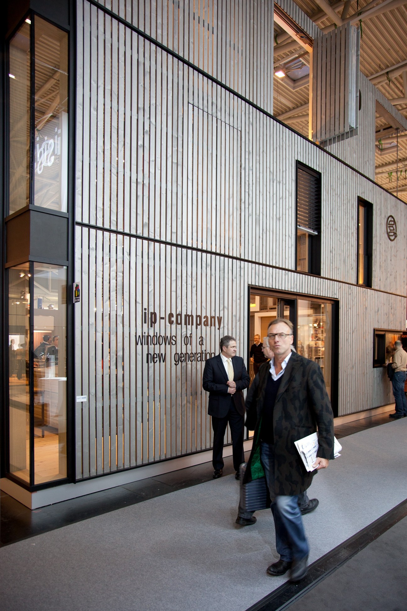 Visitors walking past a modern facade display with vertical metal slats and text reading 'ib-company windows of a new generation' at BAU exhibition.
