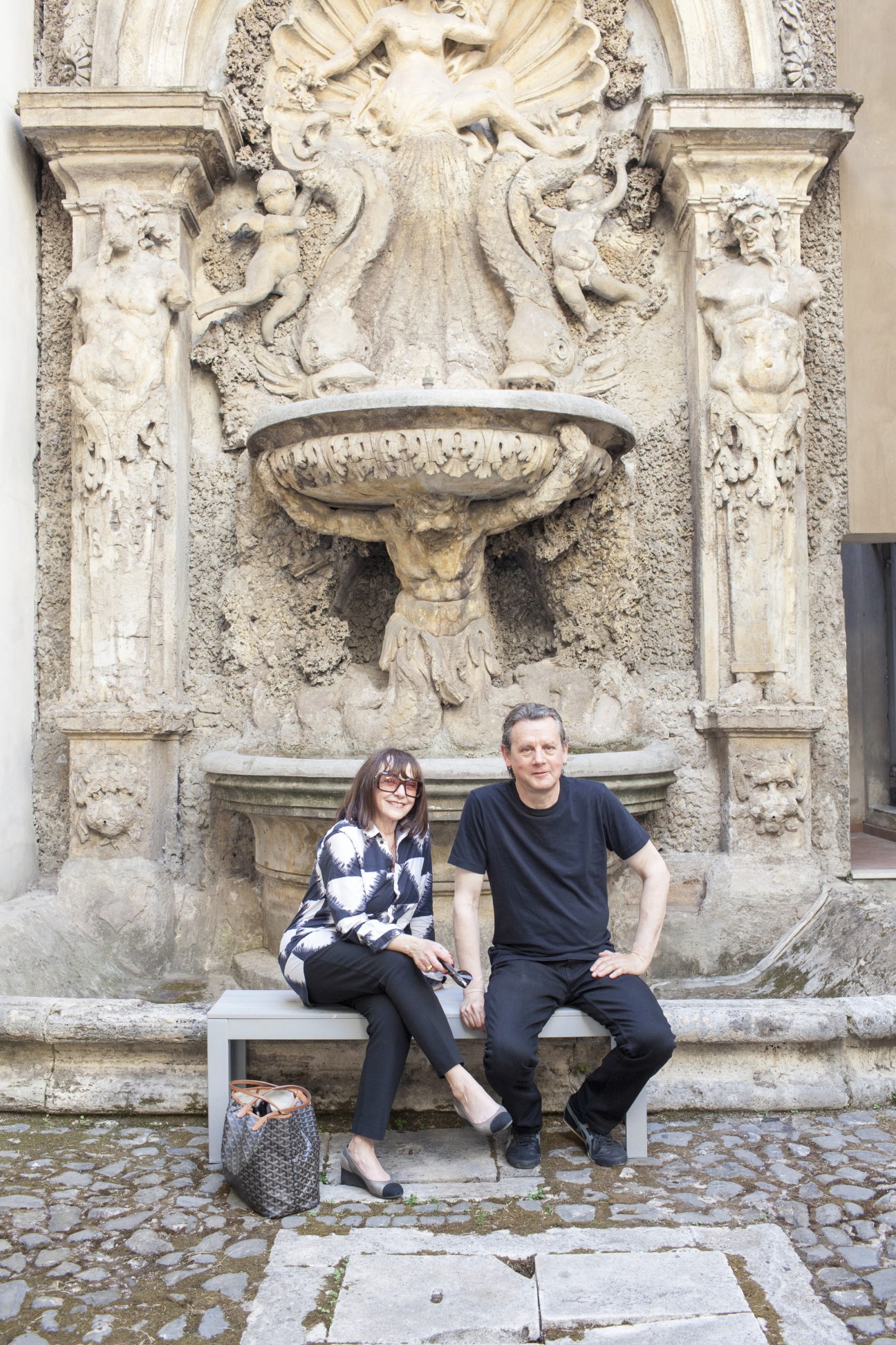 Two people sit on a white bench beneath an ornate baroque stone fountain relief in a Rome courtyard.