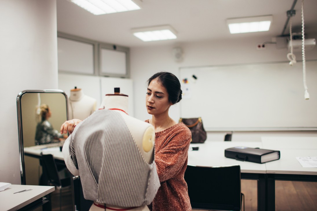 A fashion student adjusts a white pinstriped garment on a dress form in a bright studio classroom.