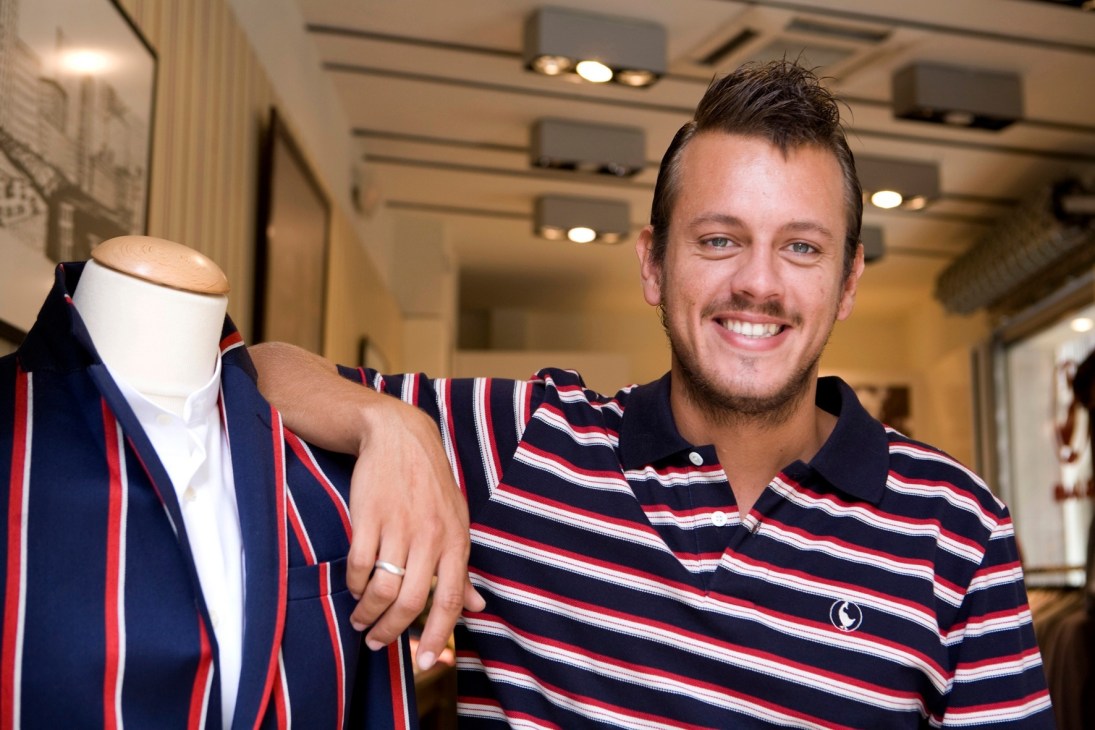 Man in striped polo shirt stands next to a dress form displaying a navy and red striped blazer in a modern retail space.