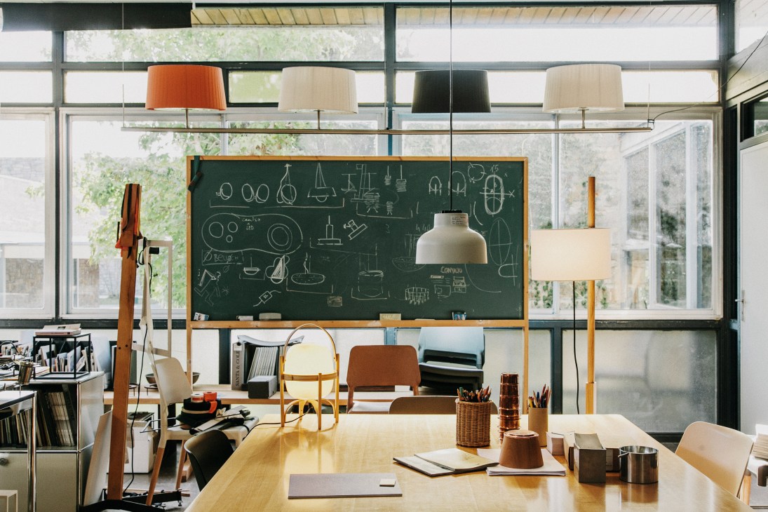 Bright modern office with large chalkboard wall, hanging pendant lamps, wooden table, and floor-to-ceiling windows overlooking greenery.
