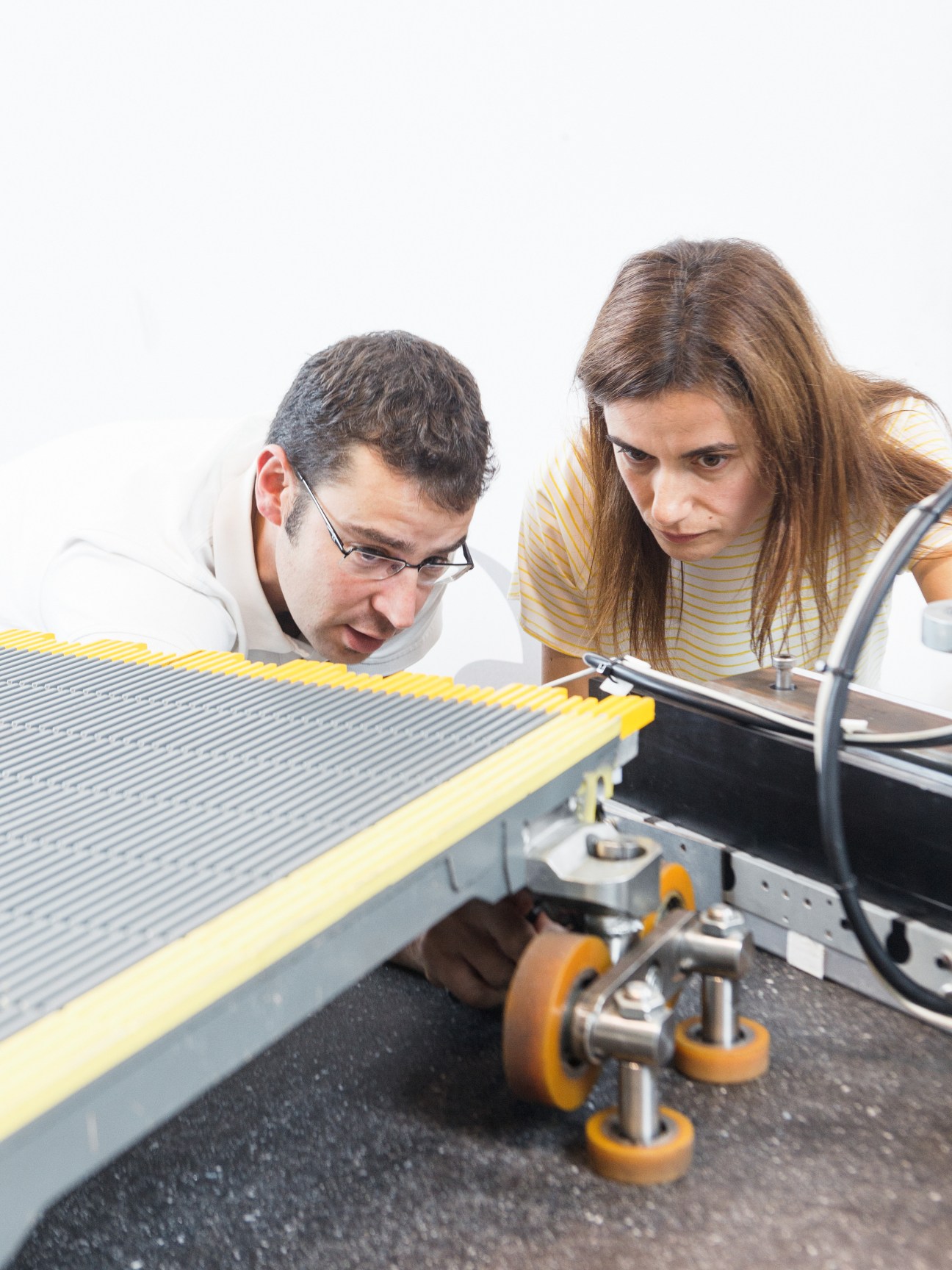 Two researchers examine a yellow and grey moving walkway model with orange wheels at ThyssenKrupp Elevator's innovation centre.