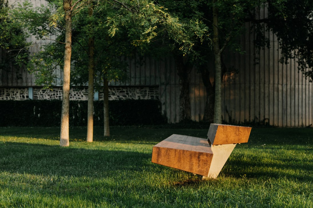 Trapecio bench with orange and natural wood panels on grass beneath trees in a garden setting.