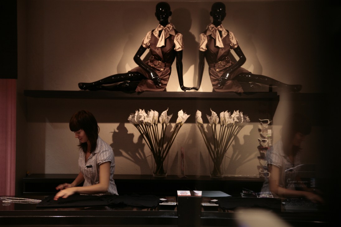 A boutique shop worker arranges a display with mannequins wearing brown dresses and cream blouses in People's Company shopping mall.