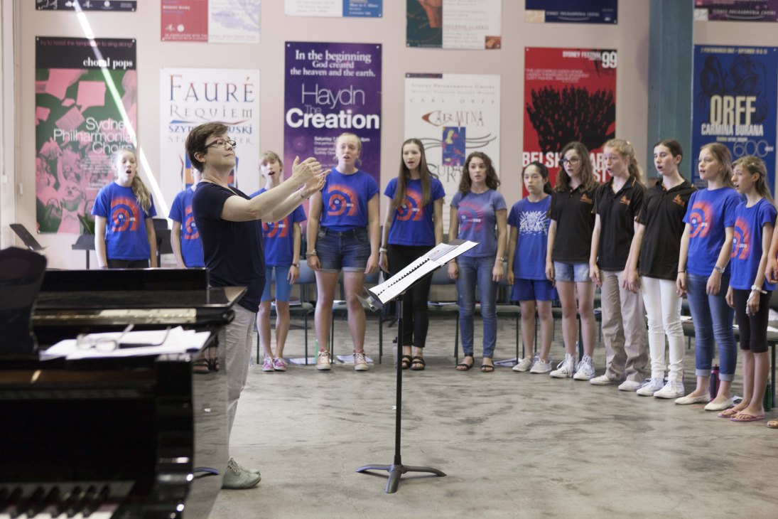 Choir director Lyn Williams leads children in blue Gondwana t-shirts during rehearsal in front of concert posters.