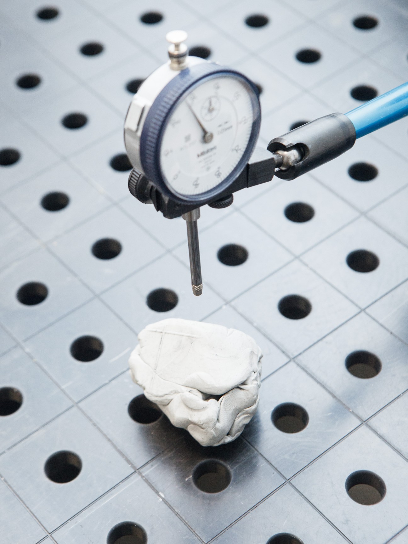 A precision dial gauge measures a white material sample on a perforated testing surface.