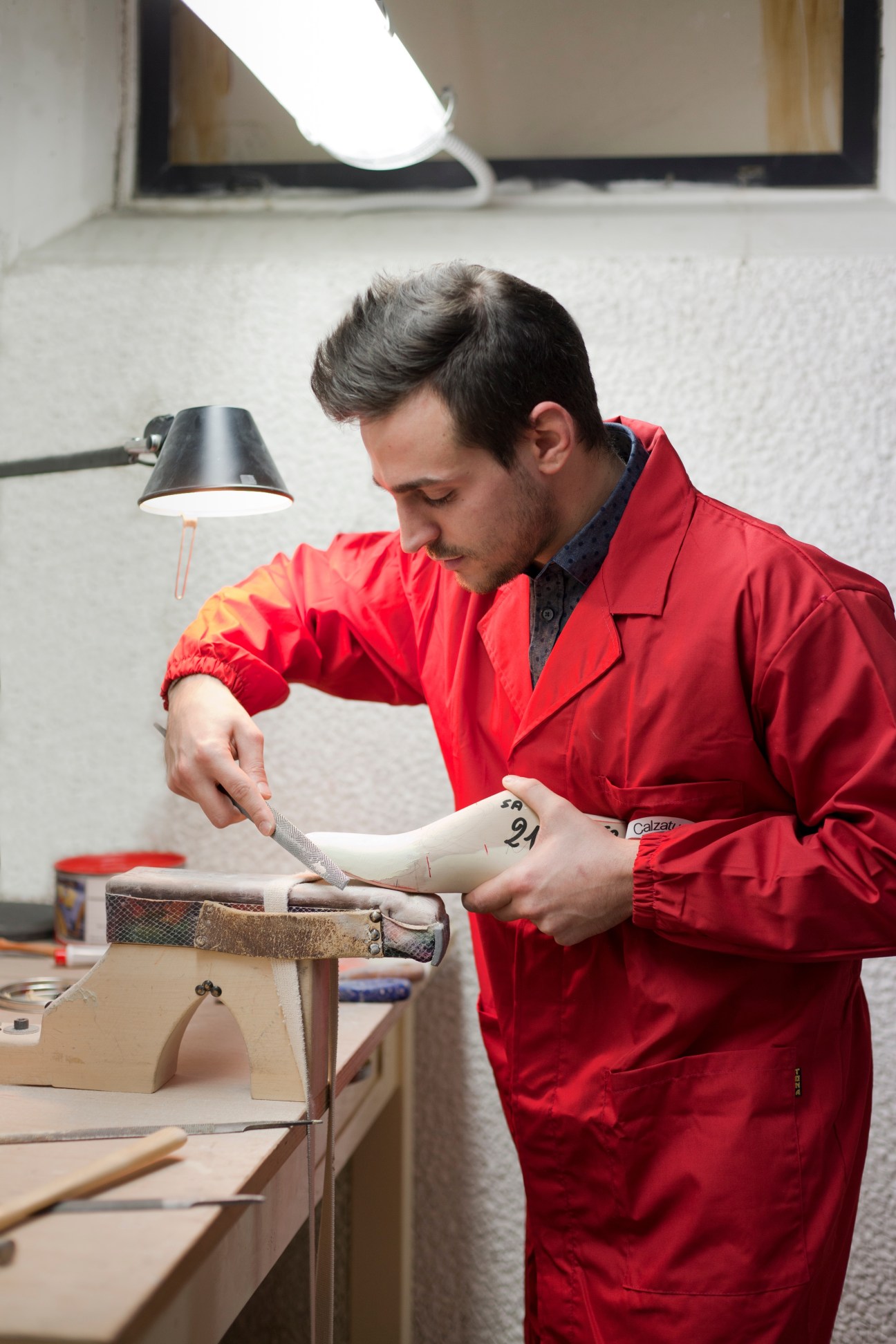 Matteo Valle shapes a wooden shoe last with a file under a desk lamp at Santoni.