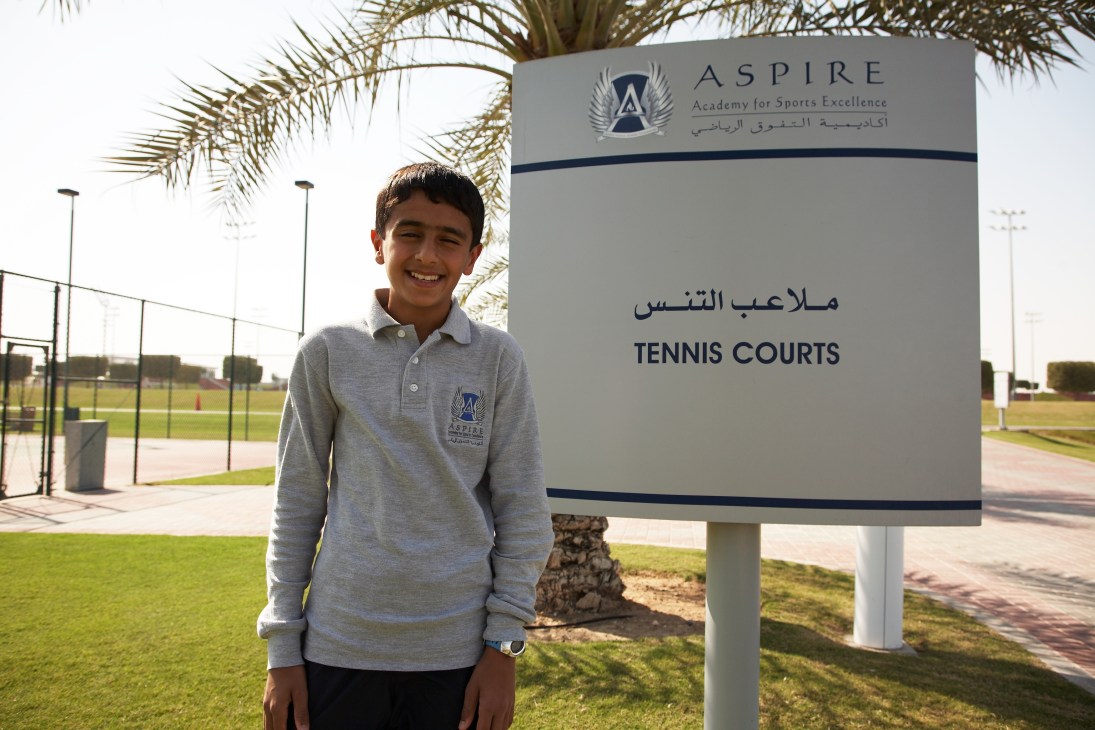 Mohammed Nasser Khinji, 13, stands smiling at an Aspire Academy tennis courts sign in Qatar.