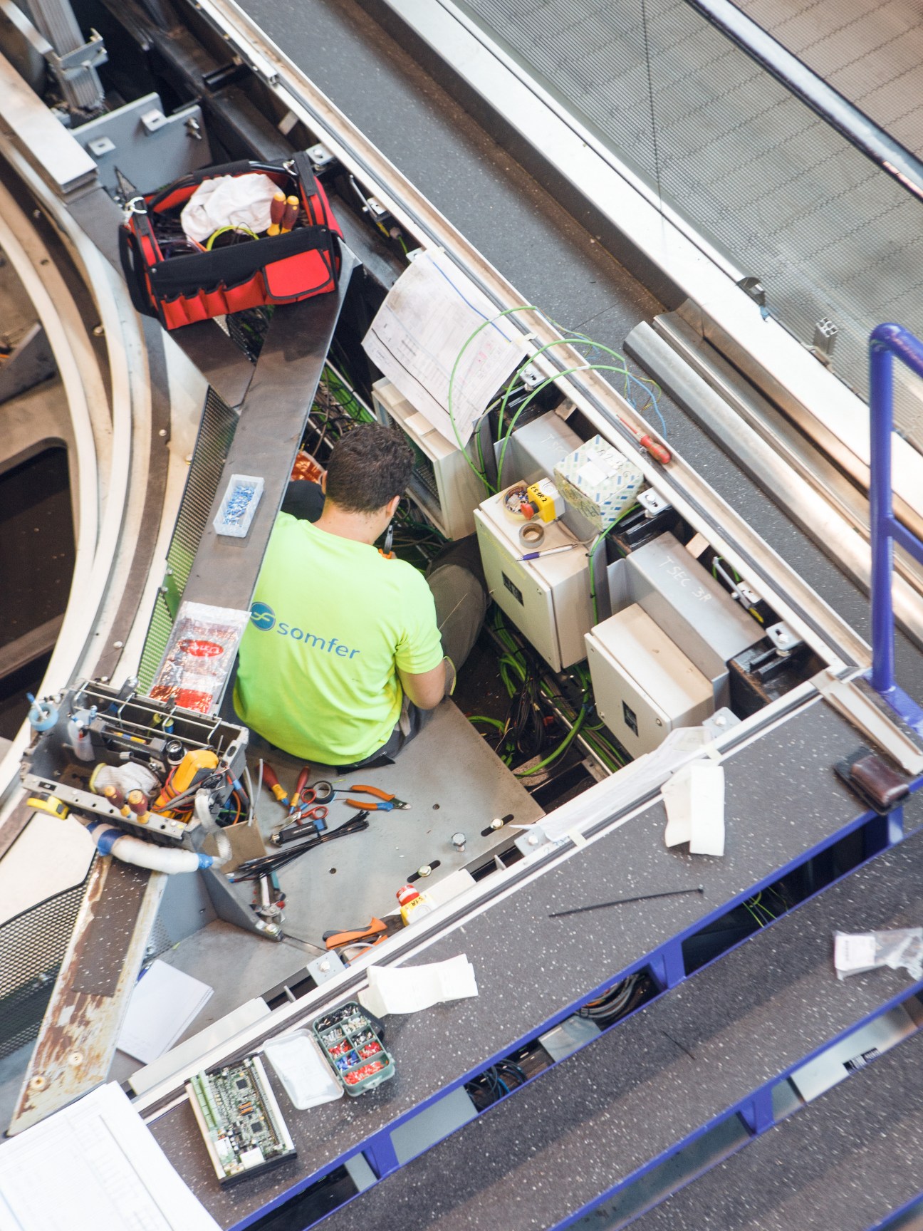 Technician in yellow safety vest working on elevator mechanical components and wiring from above.