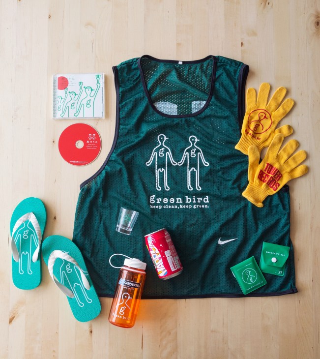 Green Bird merchandise laid out on wooden surface including Nike bib, yellow gloves, teal sneakers, water bottle and branded items.