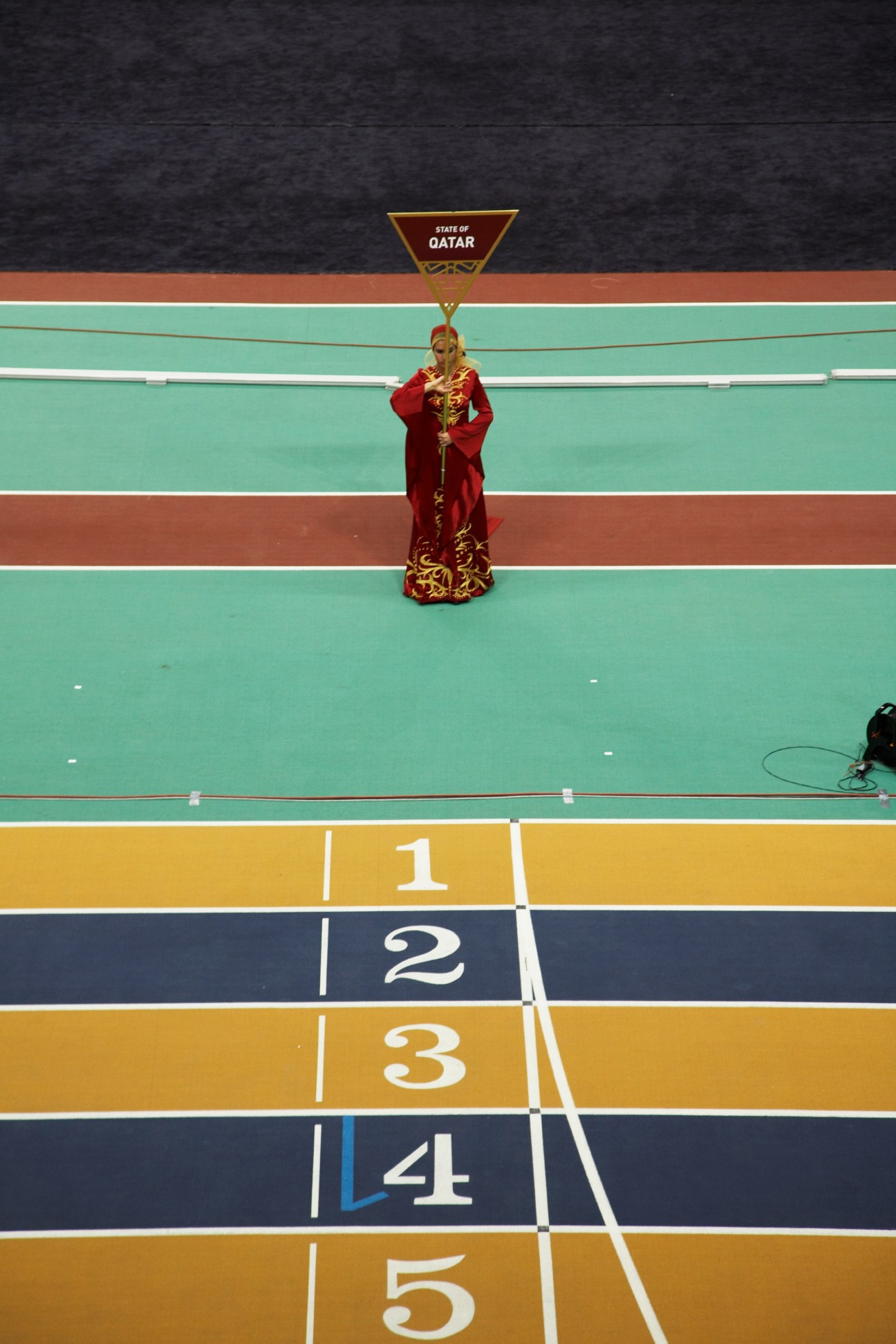 A figure in traditional Qatari dress stands on a podium with a State of Qatar banner above during an athletics championship dress rehearsal.