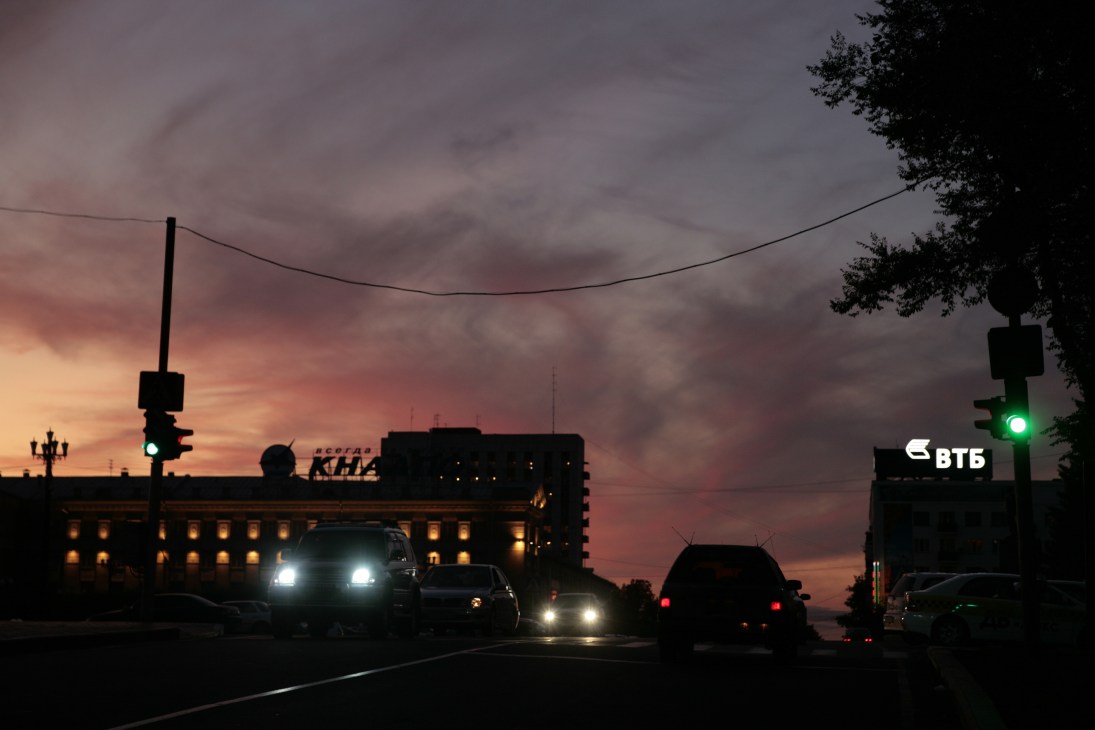 Sunset over Lenin Square in Khabarovsk with silhouetted buildings, traffic light, and cars at dusk.