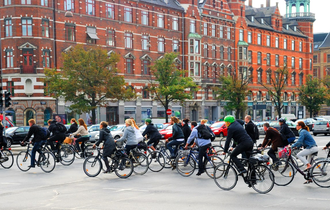 Cyclists riding on a Copenhagen street with historic red brick buildings in the background.