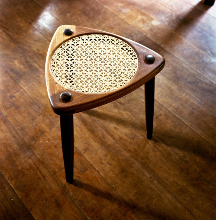 Wooden stool with woven rattan seat and three dark legs on a wood floor.