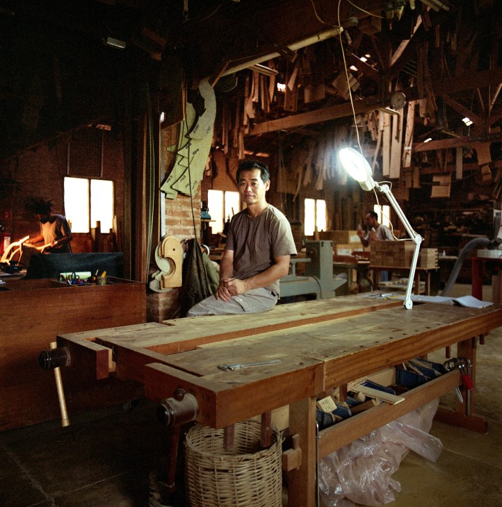Japanese carpenter Morito Ebine sits at a wooden workbench in his workshop surrounded by tools and wooden furniture pieces.