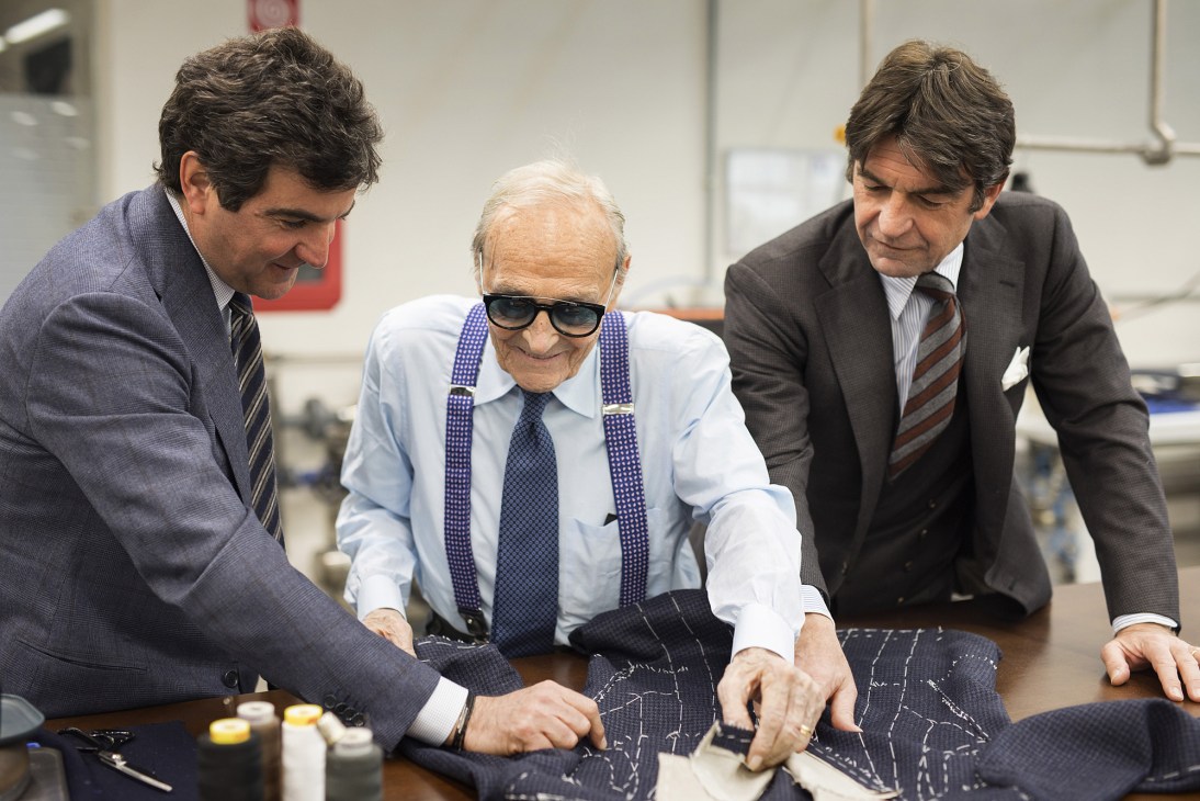 Three men in business attire examining tailored suit fabric at a workbench at Cesare Attolini.