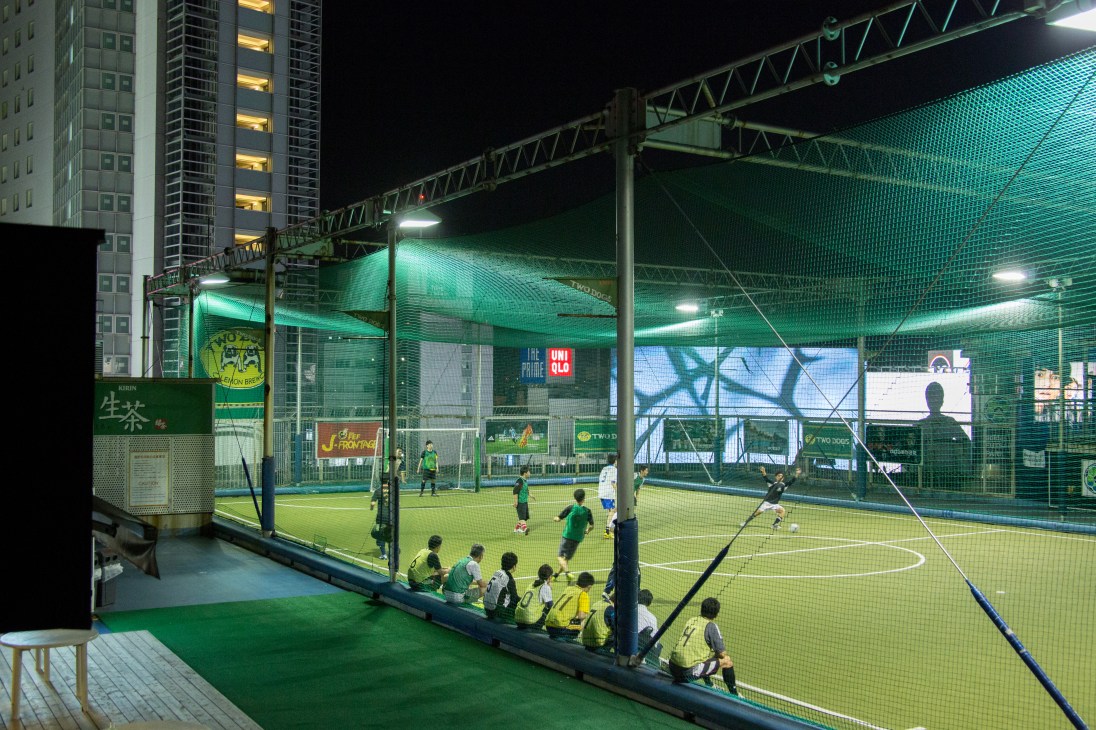 Players in yellow jerseys playing futsal on a rooftop court surrounded by green netting, with illuminated Tokyo buildings in the background at night.