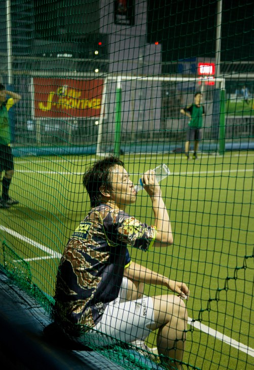 A person in athletic wear drinks water during a break from futsal at an indoor court with floodlights visible in the background.