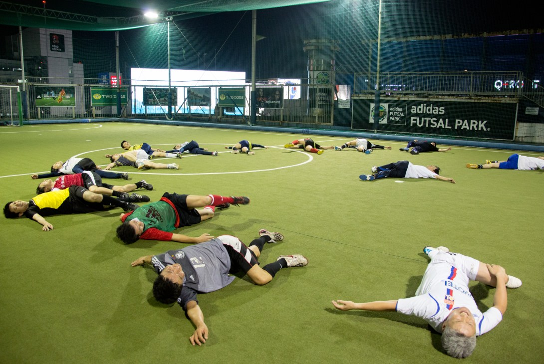 Multiple futsal players lying on artificial turf court at Adidas Futsal Park in Tokyo during cool-down exercises after practice.