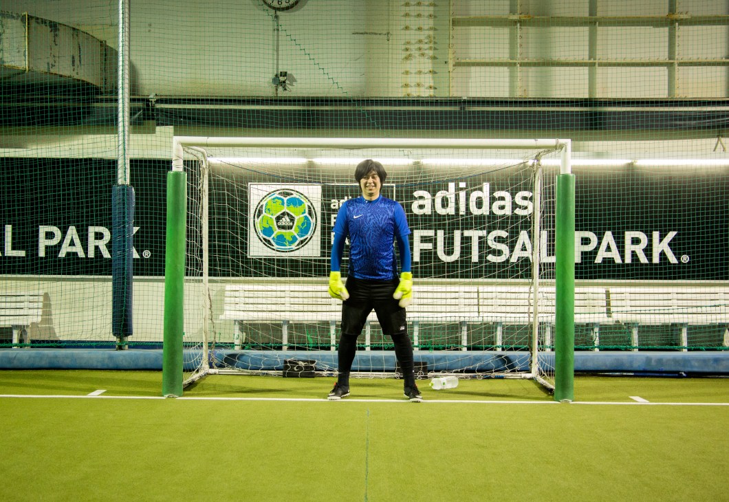 Goalkeeper in blue jersey and yellow gloves stands in goal at Adidas Futsal Park in Tokyo.