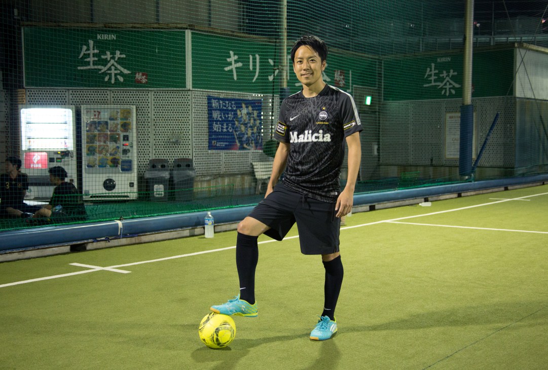 Male futsal player posing with foot on yellow ball on indoor green court with nets and advertising boards behind.