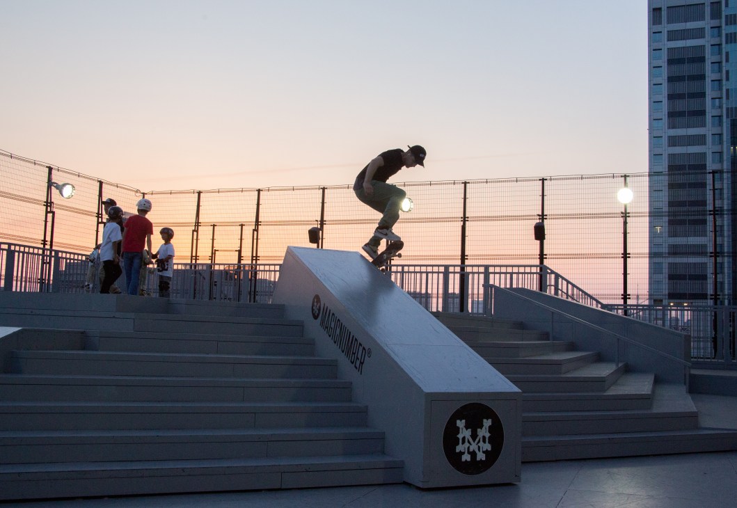 A skateboarder performs a trick on a ramp at sunset with Tokyo's urban skyline in the background.