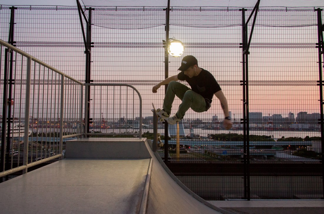 Skateboarder performing trick on rooftop ramp at dusk with city skyline.