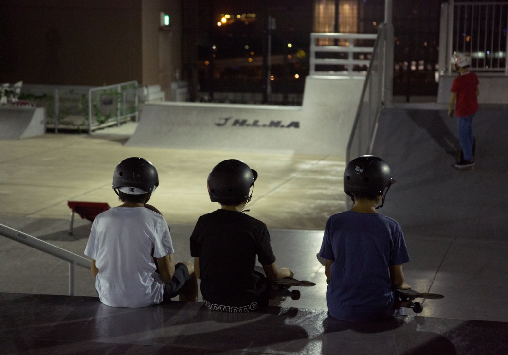 Three young skateboarders wearing helmets sit on a concrete skate park at night in Tokyo.