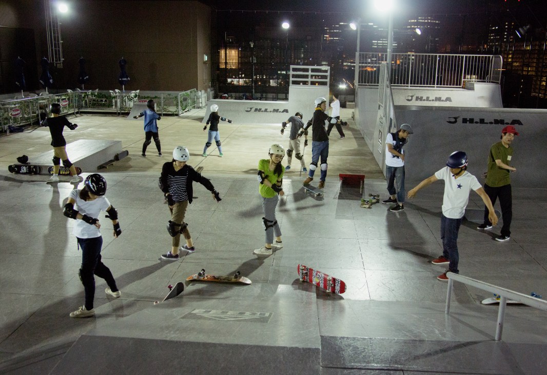 Young skateboarders practice tricks on a lit rooftop plaza at night in Tokyo.