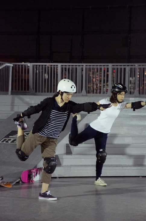Two young people skateboarding at night while balancing on one leg with arms extended.
