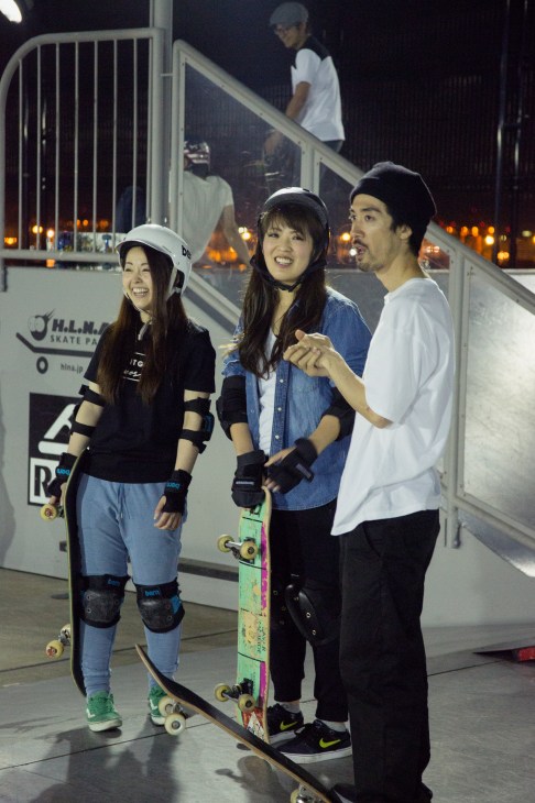 Young skateboarders in protective gear laugh together at an indoor skate park at night.